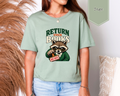 A woman wearing a sage green graphic t-shirt with a cartoon raccoon holding a 'DUE DATE!' sign and the text 'RETURN YOUR BOOKS'. The image includes a 'Sage' color swatch badge in the top right corner.