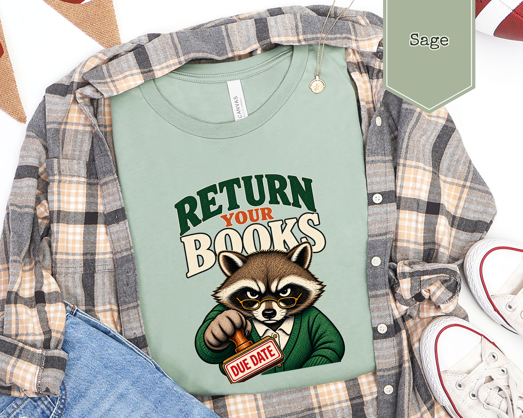 A sage green graphic t-shirt featuring a cartoon raccoon holding a 'DUE DATE' sign, styled in a flat-lay with a plaid flannel shirt, blue jeans, and white sneakers. A color swatch label in the top right indicates 'Sage'.