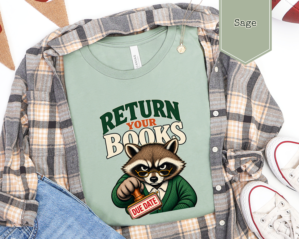 A sage green graphic t-shirt featuring a cartoon raccoon holding a 'DUE DATE' sign, styled in a flat-lay with a plaid flannel shirt, blue jeans, and white sneakers. A color swatch label in the top right indicates 'Sage'.