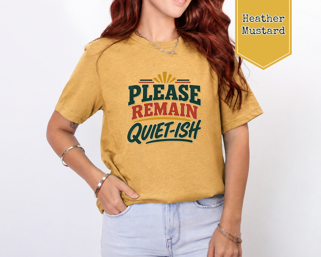 A woman wearing a heather mustard graphic t-shirt with the text 'PLEASE REMAIN QUIET-ISH' in green and red. A yellow badge in the top right corner reads 'Heather Mustard'.