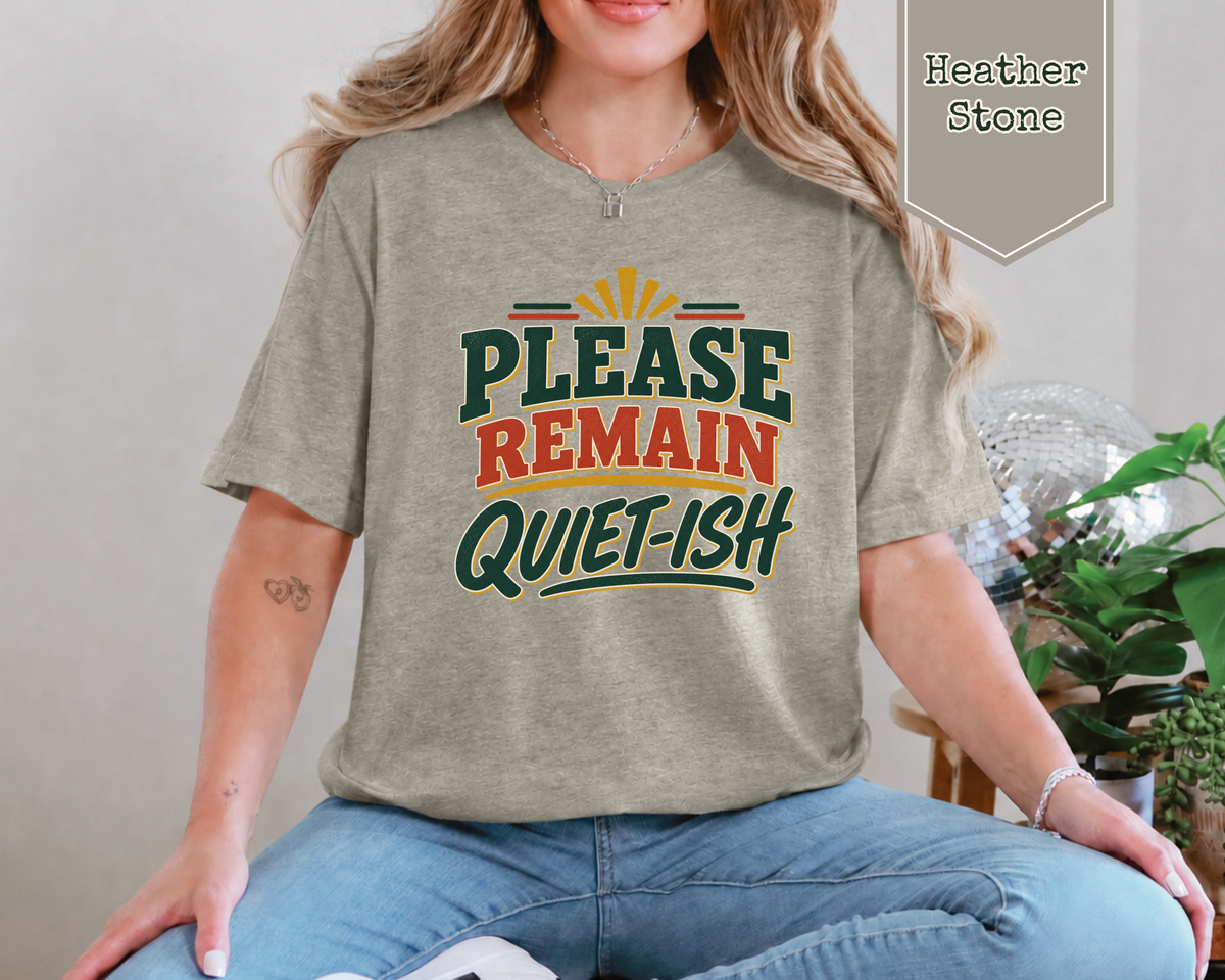 A woman wearing a heather stone graphic t-shirt that reads 'PLEASE REMAIN QUIET-ISH' in green and orange lettering, paired with blue jeans. A badge in the top right corner indicates the color is 'Heather Stone'.