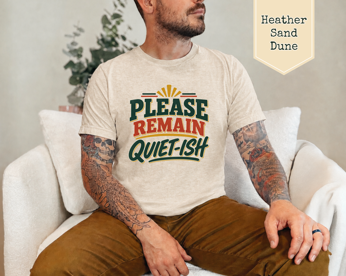 A man wearing a light heather sand t-shirt with a retro-style graphic that reads 'PLEASE REMAIN QUIET-ISH' in green and orange lettering, sitting on a white couch. A beige label in the top right corner reads 'Heather Sand Dune'.