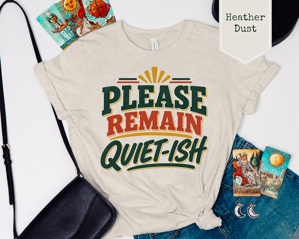 A flat-lay of a light heather dust t-shirt with a retro graphic that reads 'PLEASE REMAIN QUIET-ISH' in green, red, and yellow. The shirt is styled with blue jeans, a black crossbody bag, tarot cards, and a black hat on a white background.