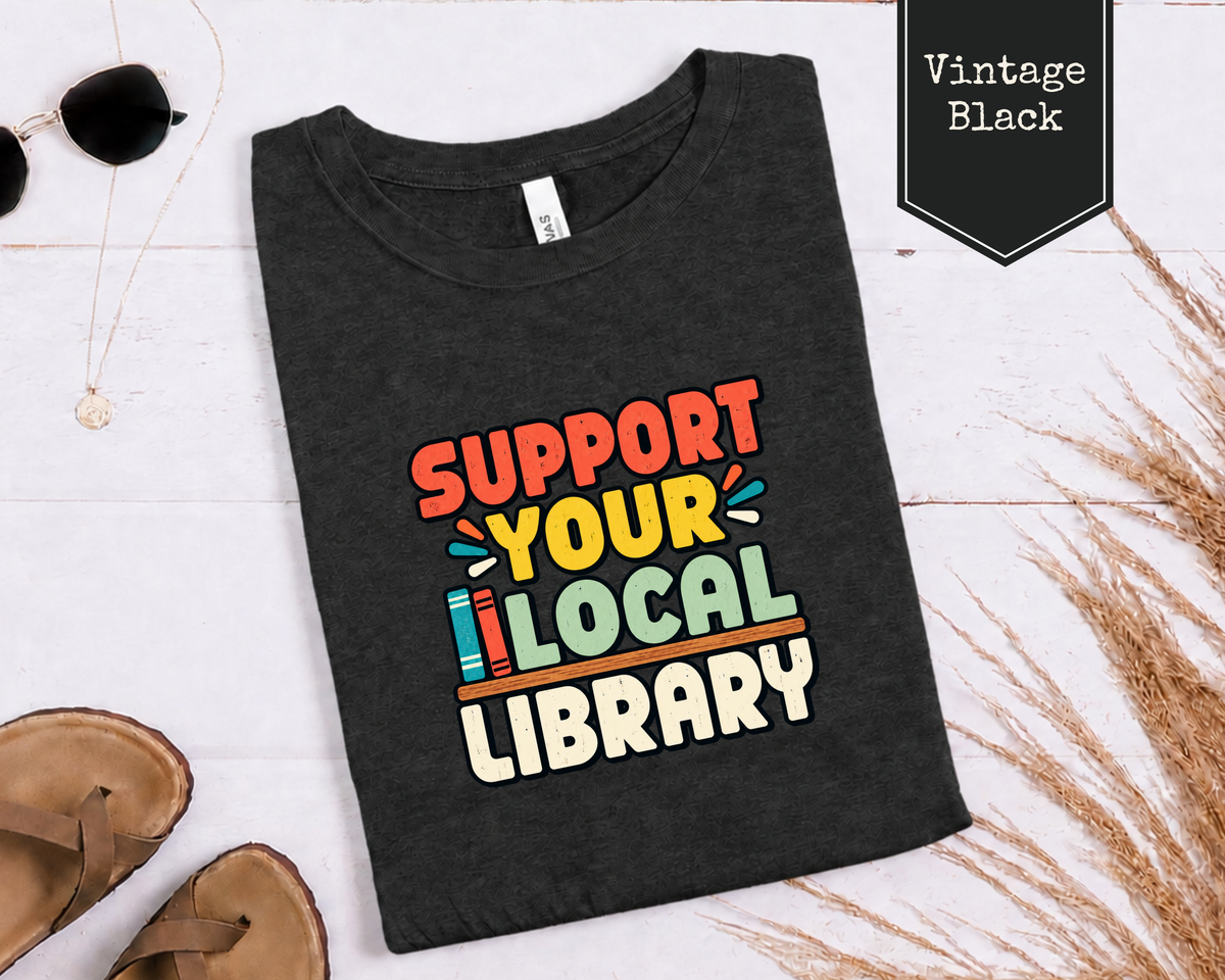 Folded Heather Dust Support Your Local Library tee styled in a flat lay with books and everyday reading accessories.