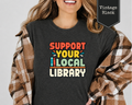 Woman wearing Heather Dust Support Your Local Library retro colorful graphic t-shirt with stacked books.