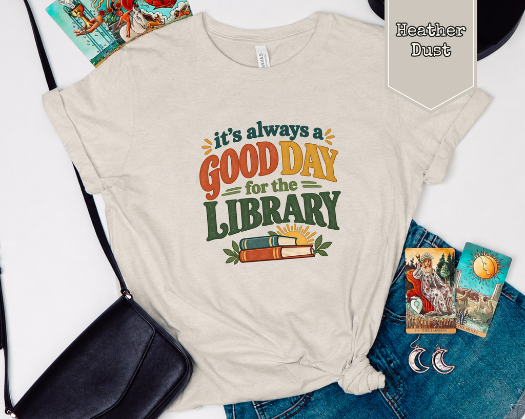 Folded Heather Dust Good Day for the Library t-shirt styled in flat lay with books, coffee mug, and accessories.