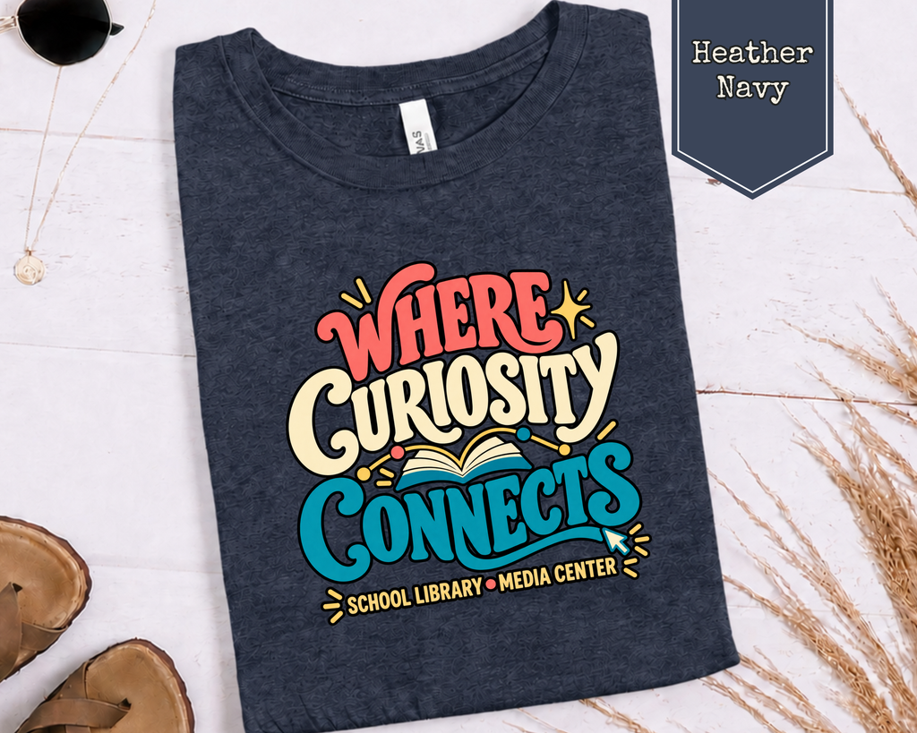 Folded Where Curiosity Connects graphic t-shirt styled in a flat lay with books, laptop, and media center accessories.
