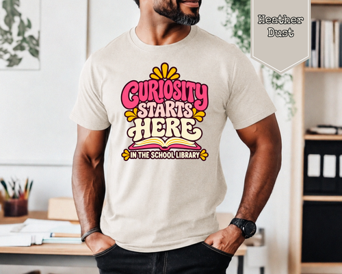 Woman wearing Heather Dust Curiosity Starts Here retro school librarian graphic tee with colorful lettering and open book artwork.