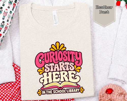 Folded Curiosity Starts Here graphic t-shirt styled in a flat lay with children's books and classroom accessories.