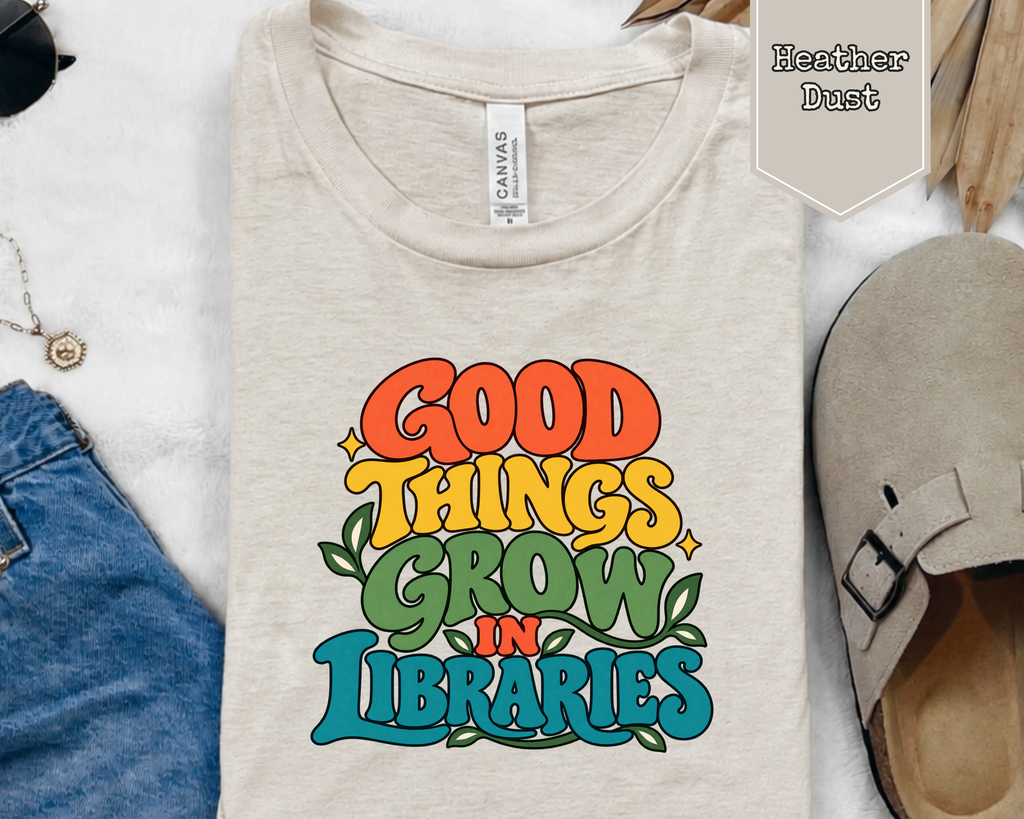 Folded Good Things Grow in Libraries graphic t-shirt styled in a flat lay with books, coffee cup, and reading accessories.