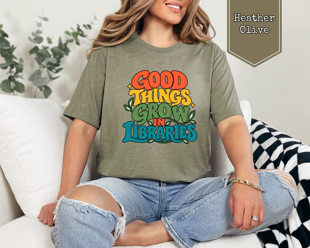 Person wearing Heather Prism Lilac Good Things Grow in Libraries unisex t-shirt in a library setting.