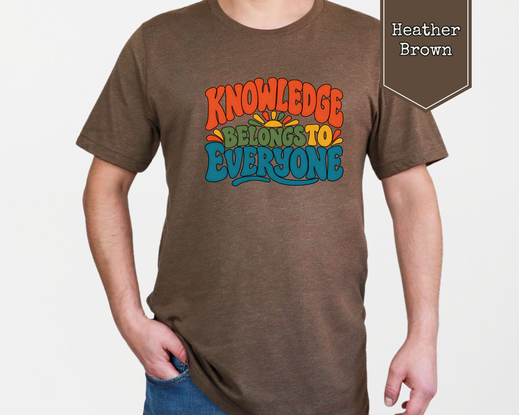 Person wearing Heather Prism Lilac Knowledge Belongs to Everyone unisex graphic t-shirt in a library setting.