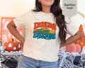 Woman wearing Heather Dust Knowledge Belongs to Everyone retro library advocacy graphic tee with colorful 1970s typography and sunrise illustration.