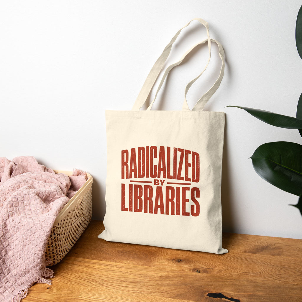 A cream canvas tote bag with the phrase 'RADICALIZED BY LIBRARIES' printed in red on the front, leaning on a wooden table against a white wall. A pink blanket sits in a woven basket to the left, and a green plant leaf is visible on the right.