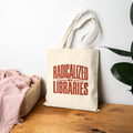 A cream canvas tote bag with the phrase 'RADICALIZED BY LIBRARIES' printed in red on the front, leaning on a wooden table against a white wall. A pink blanket sits in a woven basket to the left, and a green plant leaf is visible on the right.
