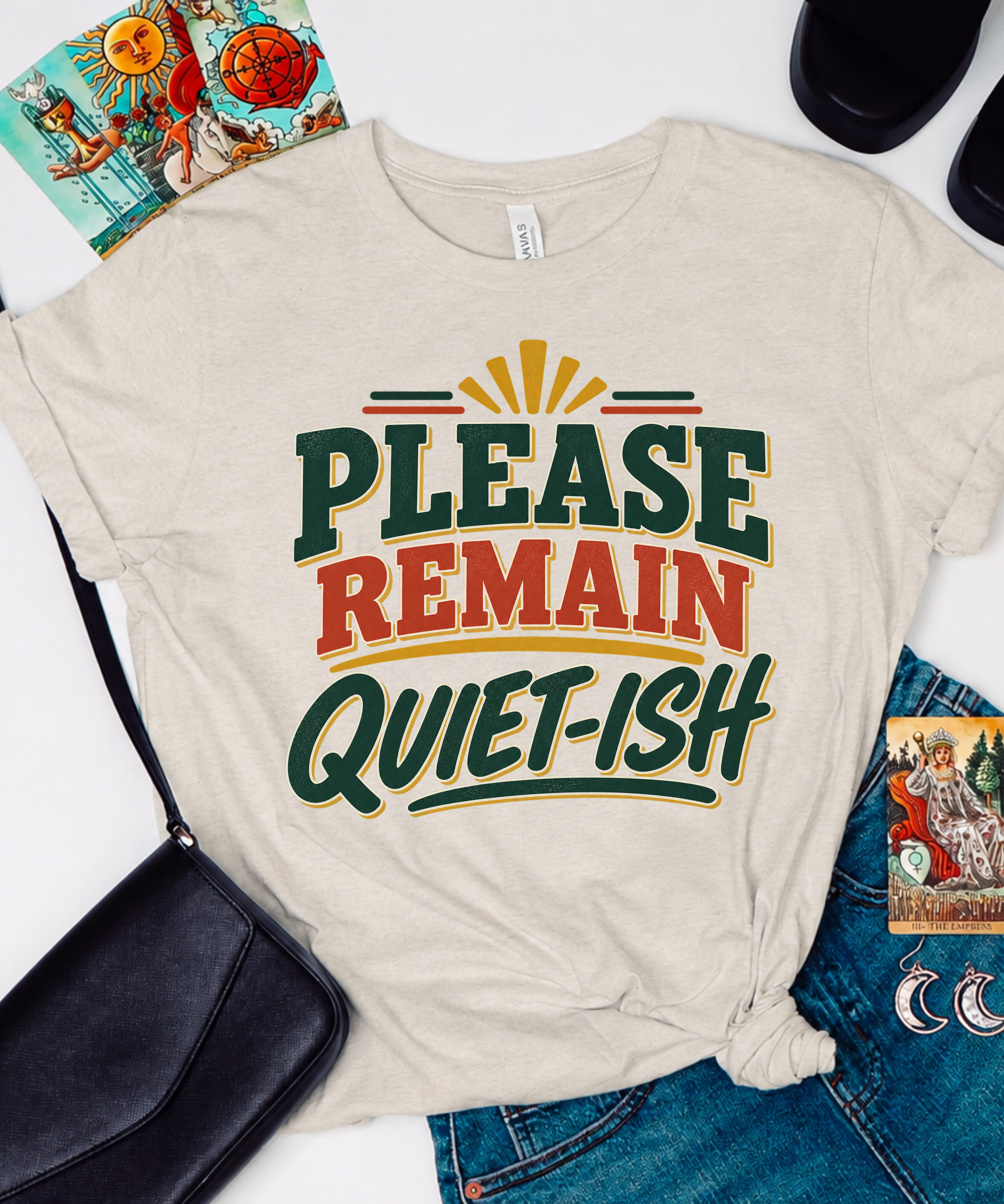 A beige graphic T-shirt laid flat with a black shoulder bag, blue jeans, and small accessories around it. The shirt has large text reading “PLEASE REMAIN QUIET-ISH” in dark green, red, and yellow tones.
